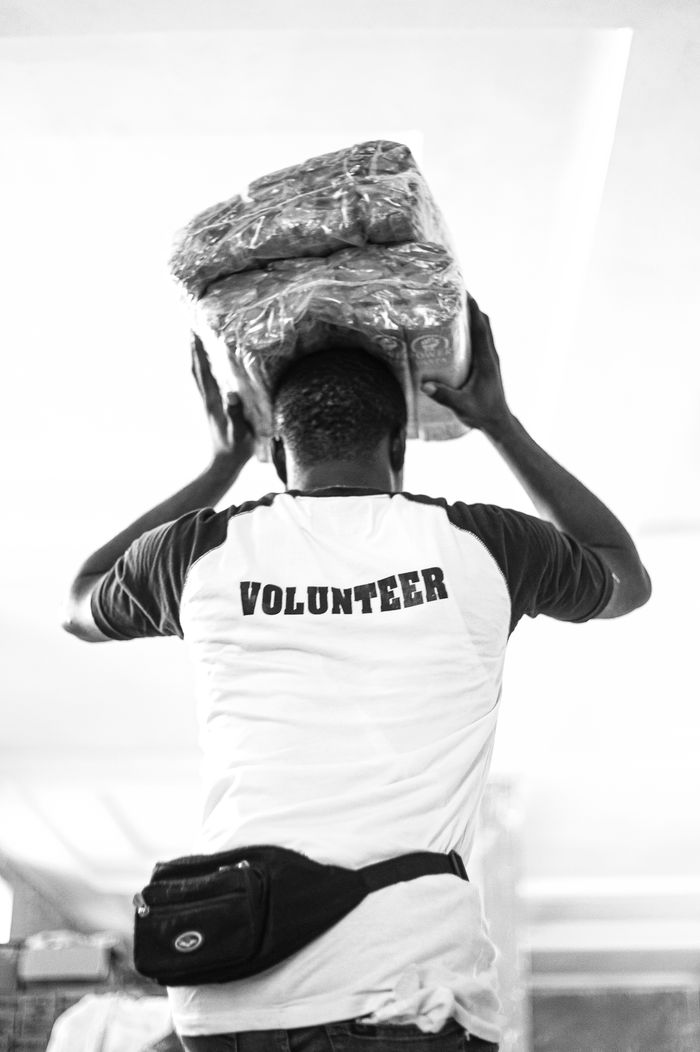 Back view of a volunteer in monochrome carrying packaged goods on their head indoors.